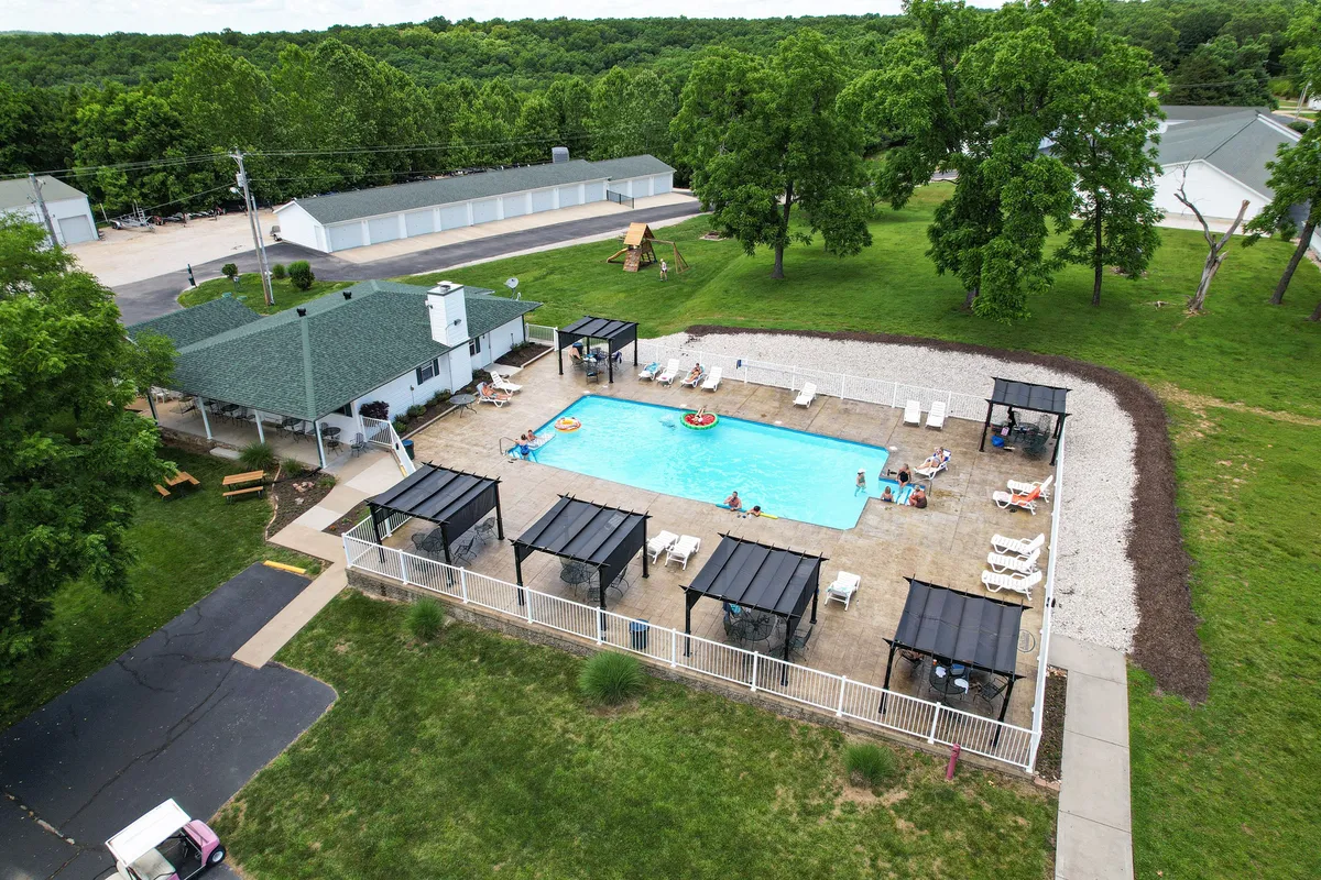 This image shows a sparkling swimming pool surrounded by poolside cabanas, lush green trees, and lawn areas. The scene is cheerful and inviting, with space for both relaxation and social gatherings. The setting suggests opportunities for both family fun and tranquil sunbathing.