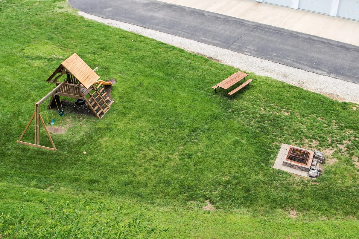 A playful, family-friendly outdoor space featuring a playground structure, a fire pit area, and a picnic bench, all set against a backdrop of trees. The area looks inviting for children and adults alike, with open sky above and cozy gathering spots.
