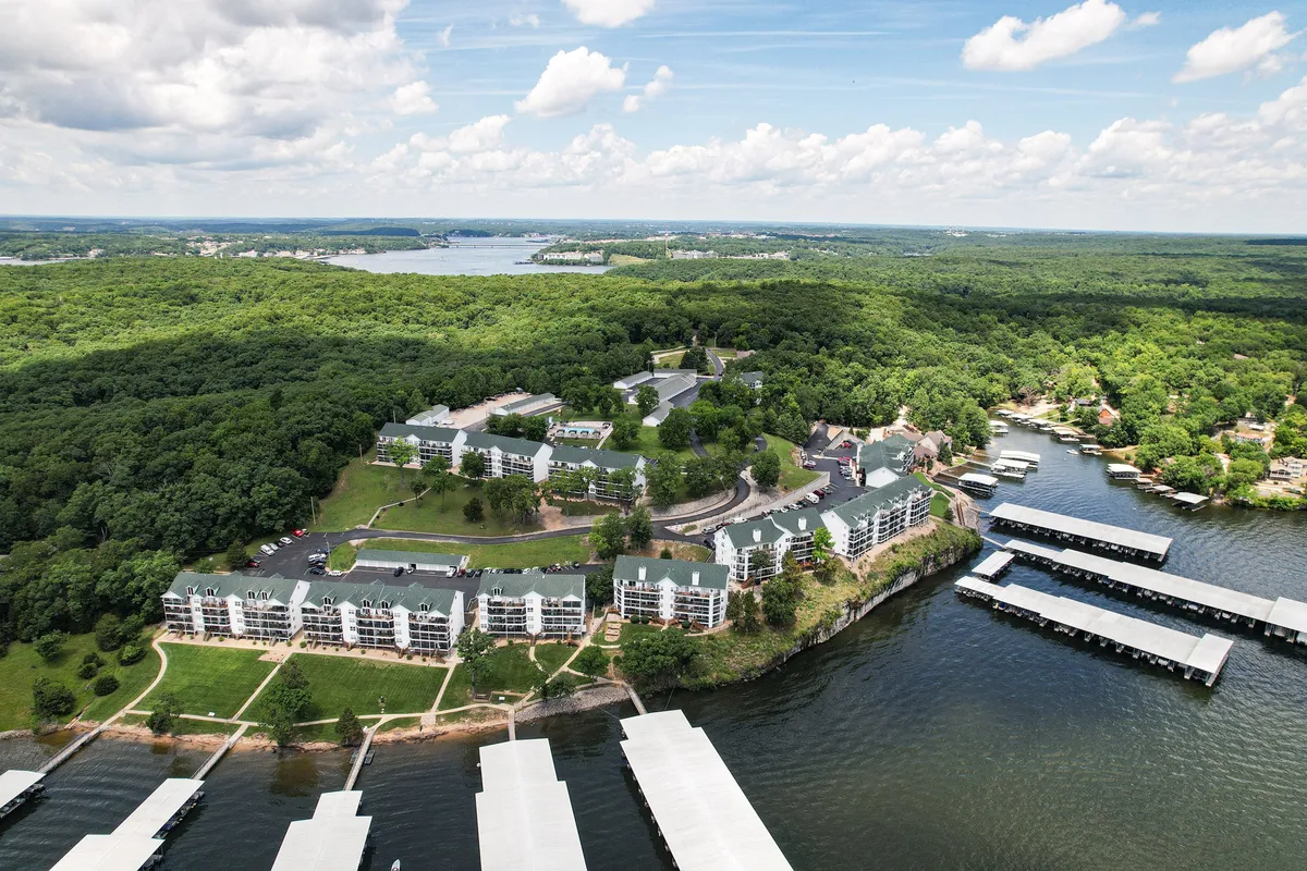 A sweeping exterior view shows a tranquil marina set against the waterfront, lush green landscape, and residential buildings. The scene is bathed in soft, natural light, suggesting either morning or evening, with boats moored and people able to walk or relax by the water.