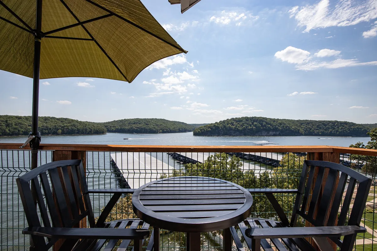 An outdoor deck setup with a wooden table and chairs under a green umbrella, overlooking a serene lake with distant hills. The scene is peaceful, inviting visitors to enjoy meals, coffee, or sunset moments outside while appreciating the lake view.