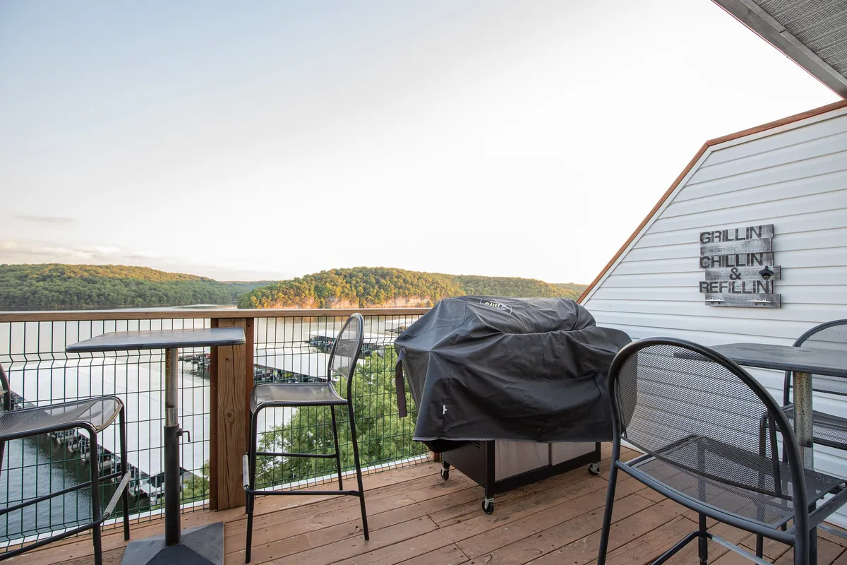 An exterior shot of a grill and outdoor seating area overlooking the water, with a sunset sky in the background. The scene exudes warmth and is set up for casual meals or gatherings with friends and family.