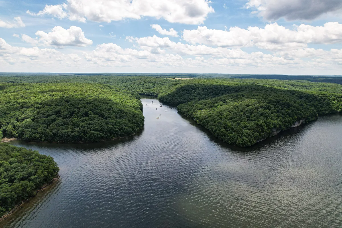 A scenic view of lush green hills, a serene river, and boats gliding across the water under a partly cloudy sky. The landscape invites exploration, with visible natural trails and wildlife potential along the riverbanks.