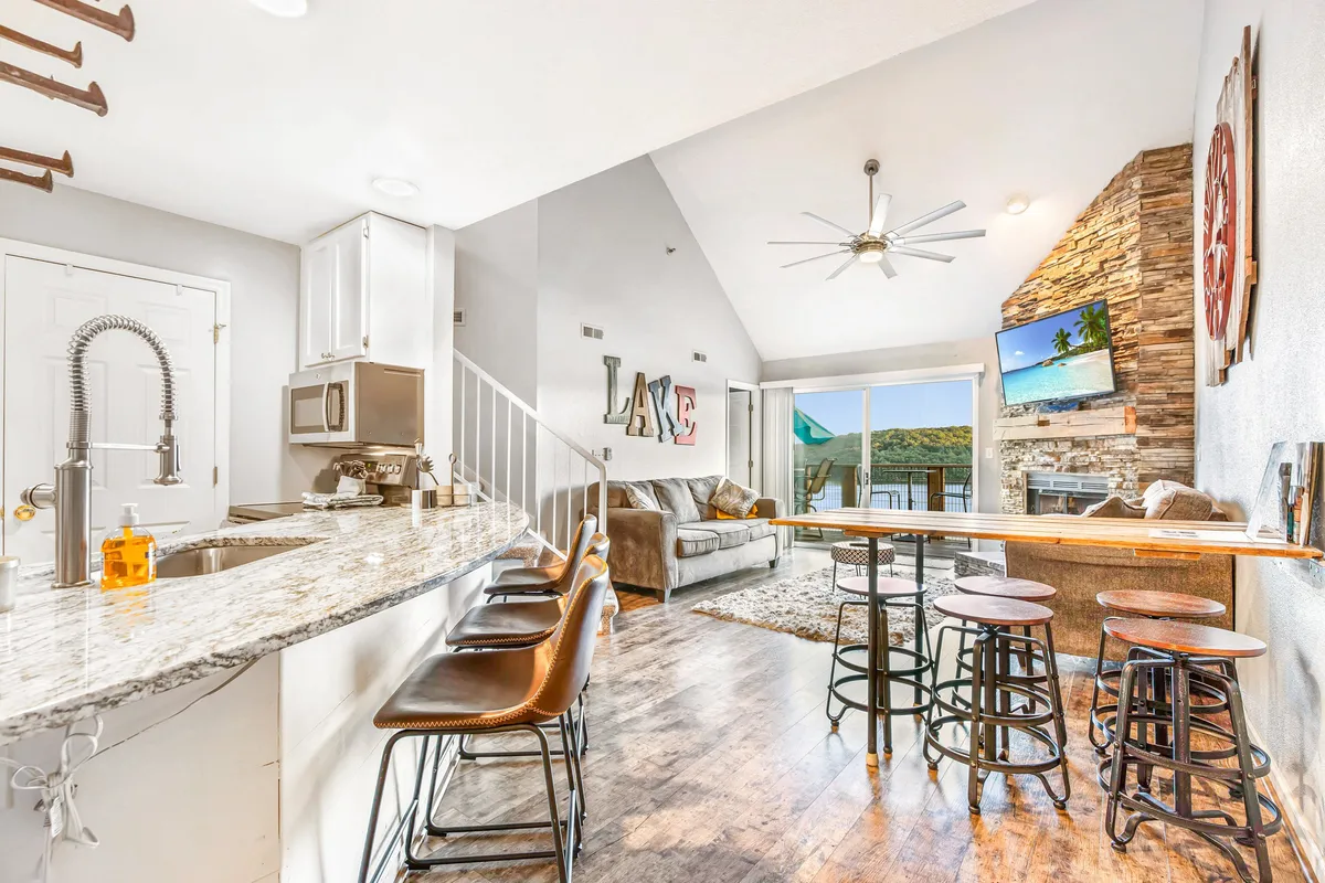A spacious, open living and kitchen area, with a granite-topped island bar and bar stools, comfortable seating, and a TV against the backdrop of large windows displaying a scenic view. The arrangement encourages both cooking and socializing, with sightlines across the room.