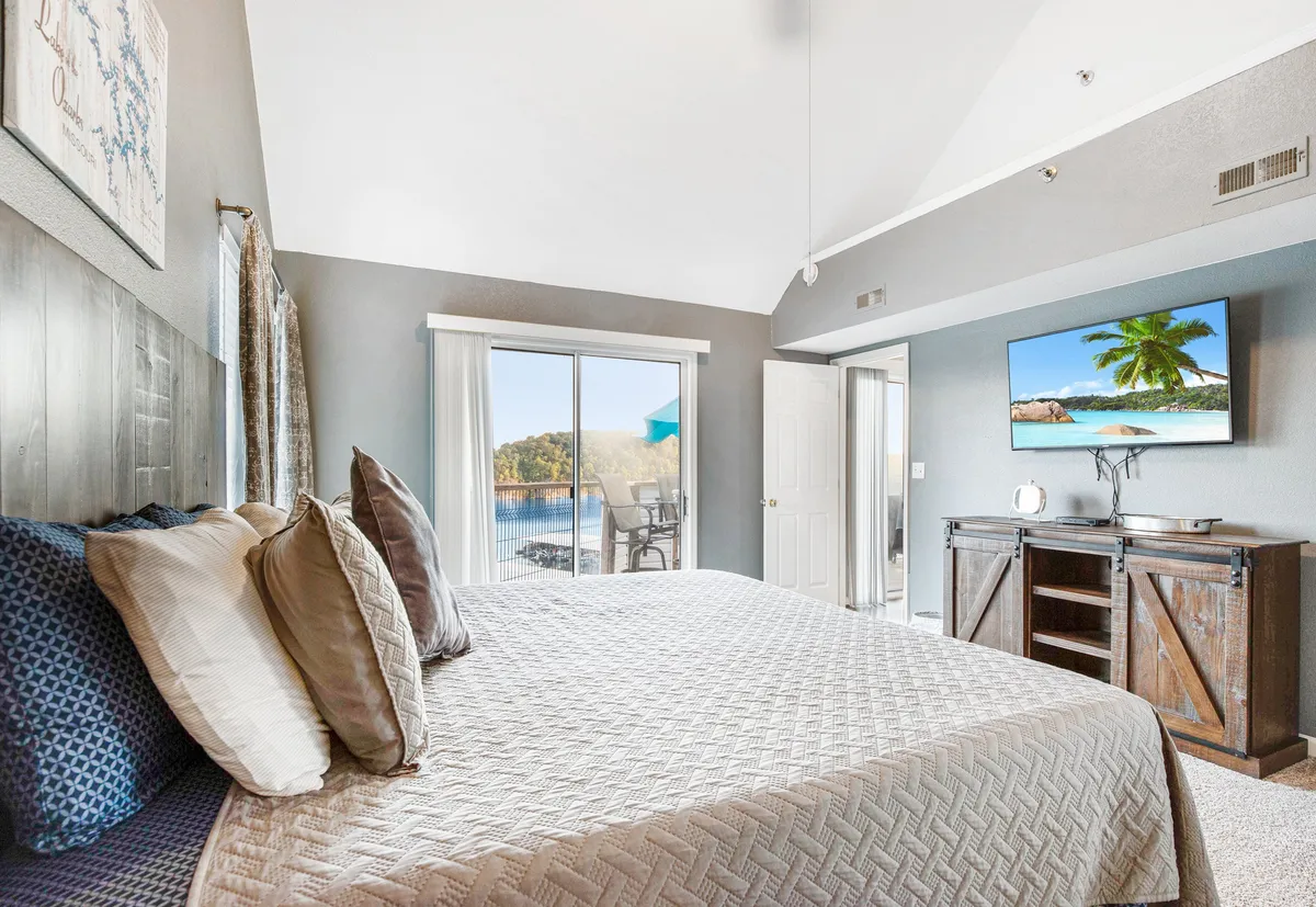 A spacious master bedroom with a large plush bed positioned to face a sliding glass door, offering sweeping views of the lake. The room also features a TV showing a beach scene, a wooden cabinet, and serene, coastal-inspired decor. Natural light fills the space, hinting at direct access to an outdoor deck.