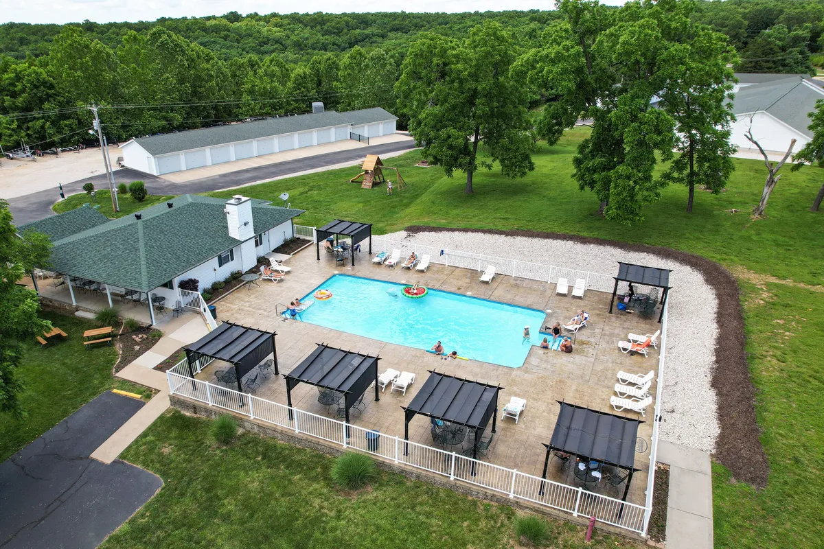 This image features the gated community swimming pool surrounded by green trees, covered seating areas, and a playground in the background. Several people are enjoying an afternoon by the pool, with children playing and others lounging or socializing nearby.