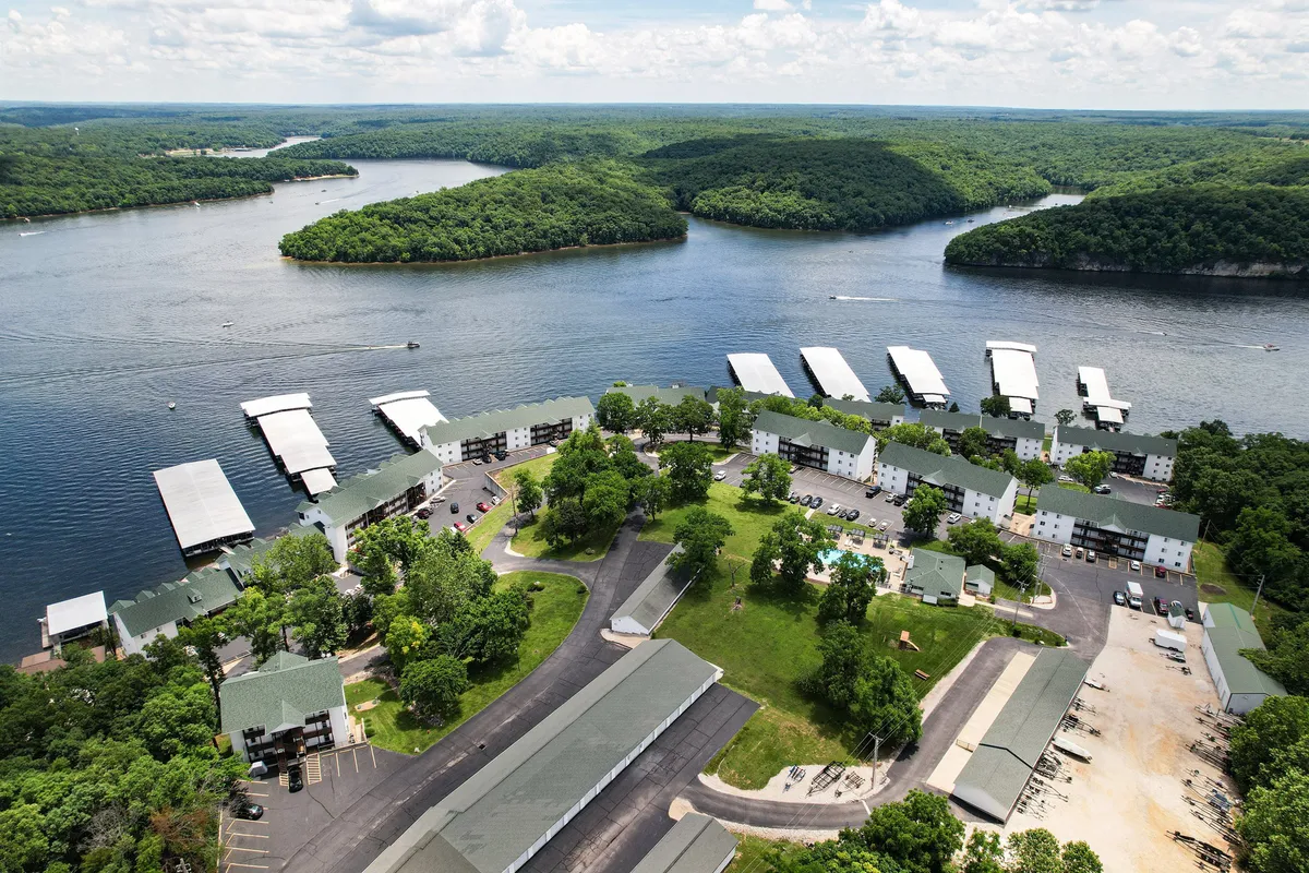 A panoramic exterior shot capturing lakeside cabins nestled among lush greenery, with a marina, sparkling swimming pool, and walking trails visible. The lake is calm, and the entire scene is bathed in warm, natural light, offering a view that feels welcoming in any season.