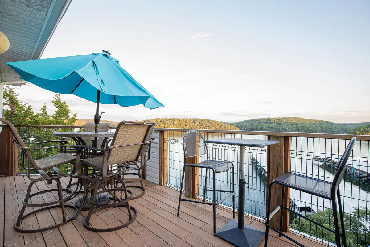 The image showcases an inviting outdoor dining area beneath a vibrant blue umbrella, set on a deck overlooking the tranquil waters of Lake of the Ozarks. The table is ready for guests, hinting at the perfect spot to enjoy meals, socialize, or unwind with a drink while soaking up breathtaking views and sunsets. The peaceful lakefront setting is complemented by the lush greenery of the state park in the background, enhancing the tranquil and exclusive atmosphere.